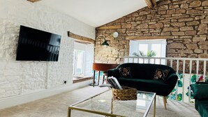 Un salon avec un mur en pierre, un canapé vert, une table basse en verre et un téléviseur fixé au mur.