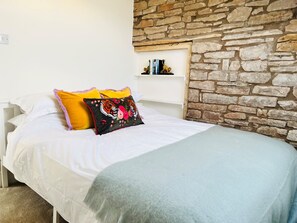 A bed with white bedding, a black pillow with a floral design, and a yellow pillow. There is a stone wall behind the bed and a shelf with books above it. - Stylish & Rural Luxury Cottage (Welsh Newton Common)