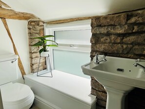 A bathroom with a white toilet, a bathtub, and a sink. There is a window above the bathtub and a plant on a stool next to the toilet. - Stylish & Rural Luxury Cottage (Welsh Newton Common)