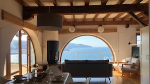 Un coin repas avec une table dressée pour deux personnes, un canapé et un téléviseur, dans une pièce dotée de poutres en bois et de grandes fenêtres en arc de cercle donnant sur la mer.