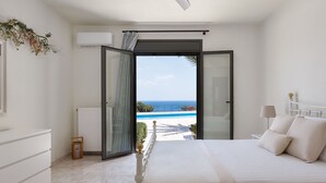 A bedroom with a bed, a nightstand, a lamp, and a view of the sea through an open door.