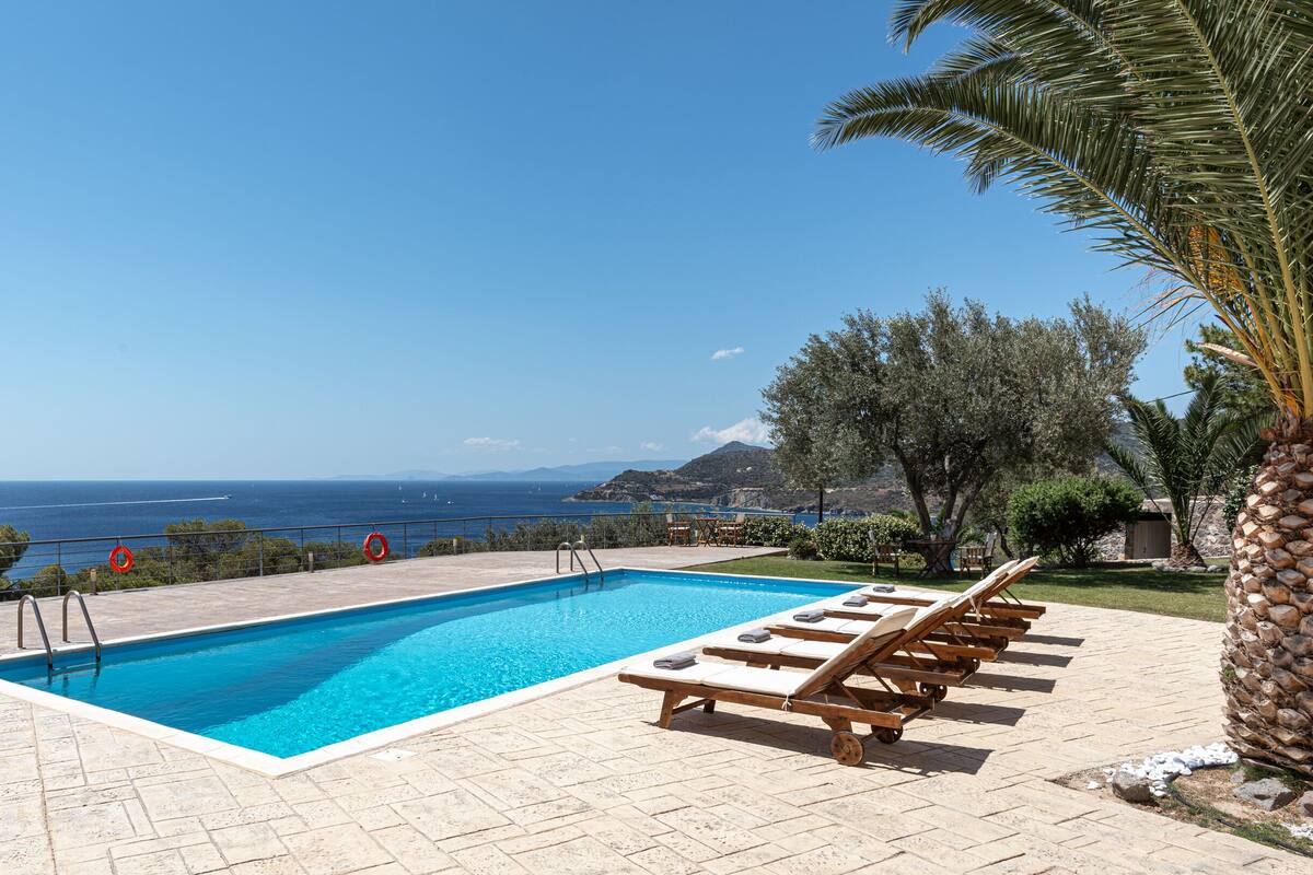Une piscine rectangulaire à l’eau d’un bleu limpide, entourée d’une terrasse dallée. Des chaises longues en bois sont alignées le long du bord de la piscine. La piscine se trouve devant un ciel d’un bleu éclatant et offre une vue lointaine sur la mer.