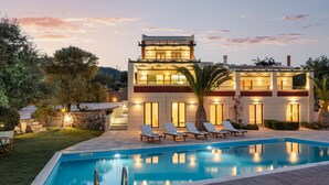 A modern two-story house with a swimming pool, lounge chairs, and a well-lit garden at dusk.