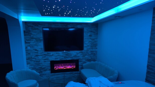 A modern room with a stone wall, a flat-screen TV, a fireplace, a sofa, and a bathtub with a blue light. - Sandspa 84 (Orange)