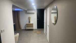 A hallway with a white wall, a round mirror, a small table with a vase, and an air conditioning unit. - Sandspa 84 (Orange)