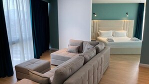 A modern hotel room with a large beige sectional sofa, a separate sofa, and a bed with white linens. - Granda Hotel (Kashar)