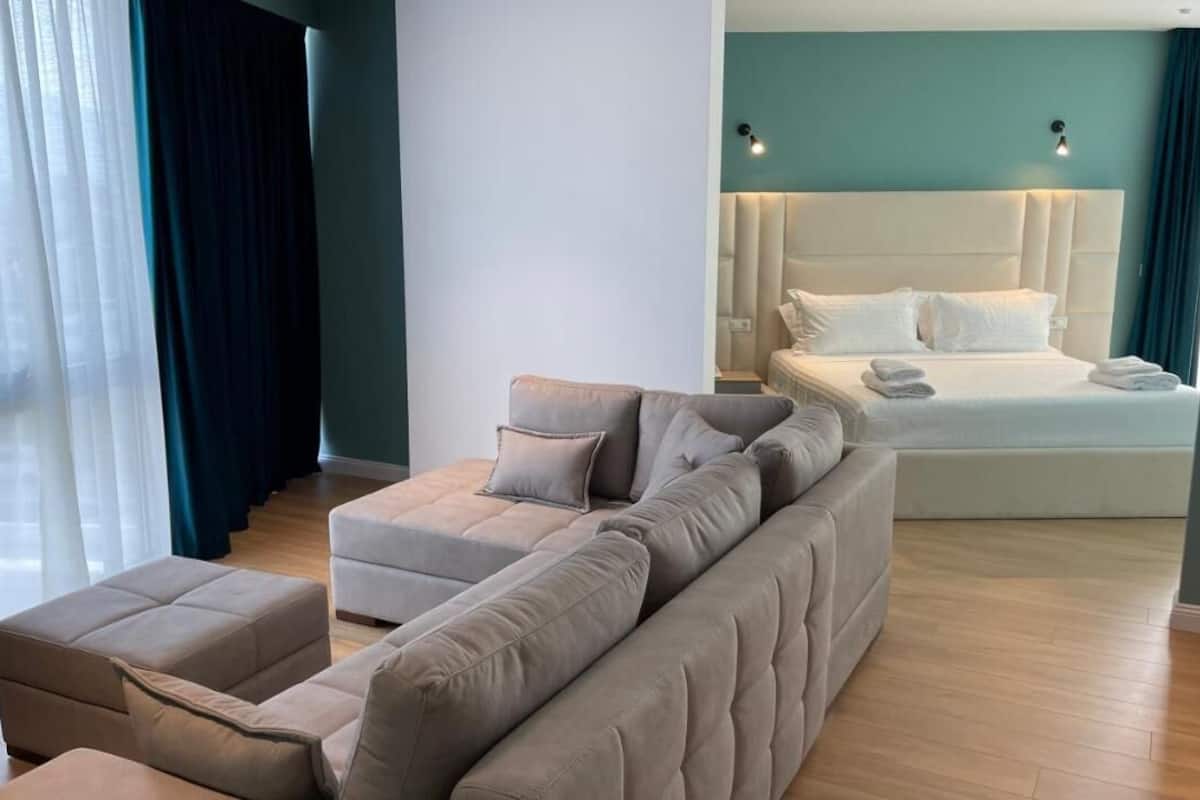 Une chambre d’hôtel moderne avec un grand canapé d’angle beige, un canapé séparé et un lit avec du linge de lit blanc.