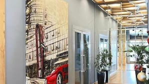 A hallway with a wooden ceiling, a large painting of a car on a building, glass doors, and potted plants. - Granda Hotel (Kashar)