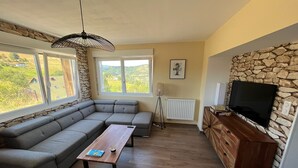 A modern living room with a sectional sofa, a wooden coffee table, a TV mounted on a stone wall, and large windows offering a view of the outdoors.