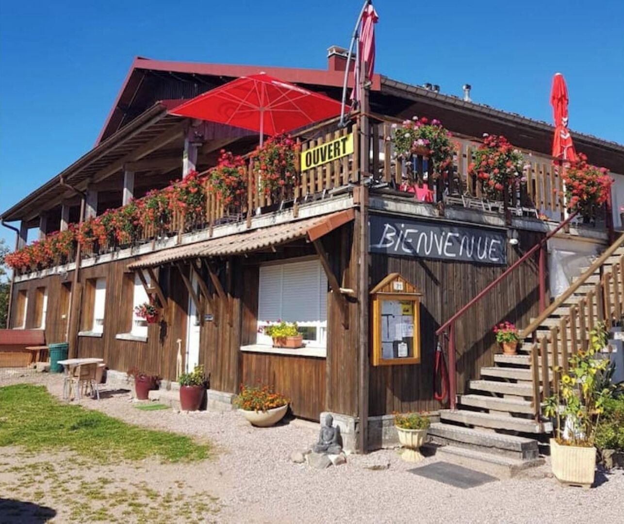 A wooden building with a balcony adorned with flowers, a sign reading "OUVERT," and a staircase leading to the entrance.