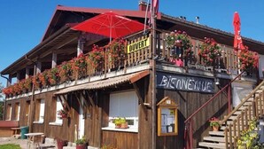 A wooden building with a balcony adorned with flowers, a sign reading "OUVERT," and a staircase leading to the entrance.