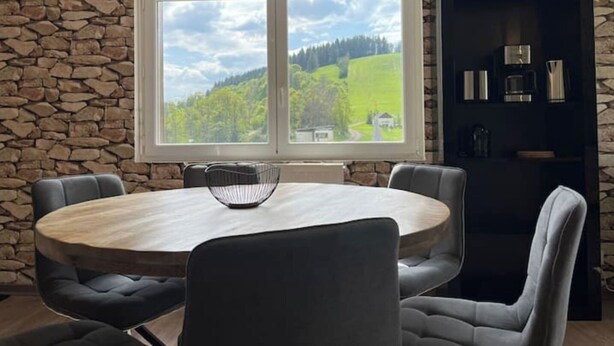 A dining area with a round table and six chairs, a stone wall, a large window with a view of greenery, and a black cabinet with items on the right.