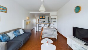 A modern living room with a sofa, a dining table with chairs, a TV, and a kitchen area in the background. - 200m from the beach, flat for 4 (Saint-Hilaire-de-Riez)