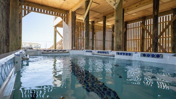 Ein Pool mit Holzträgern und einer gefliesten Kante, umgeben von einem überdachten Bereich mit Blick auf ein Gebäude im Freien.