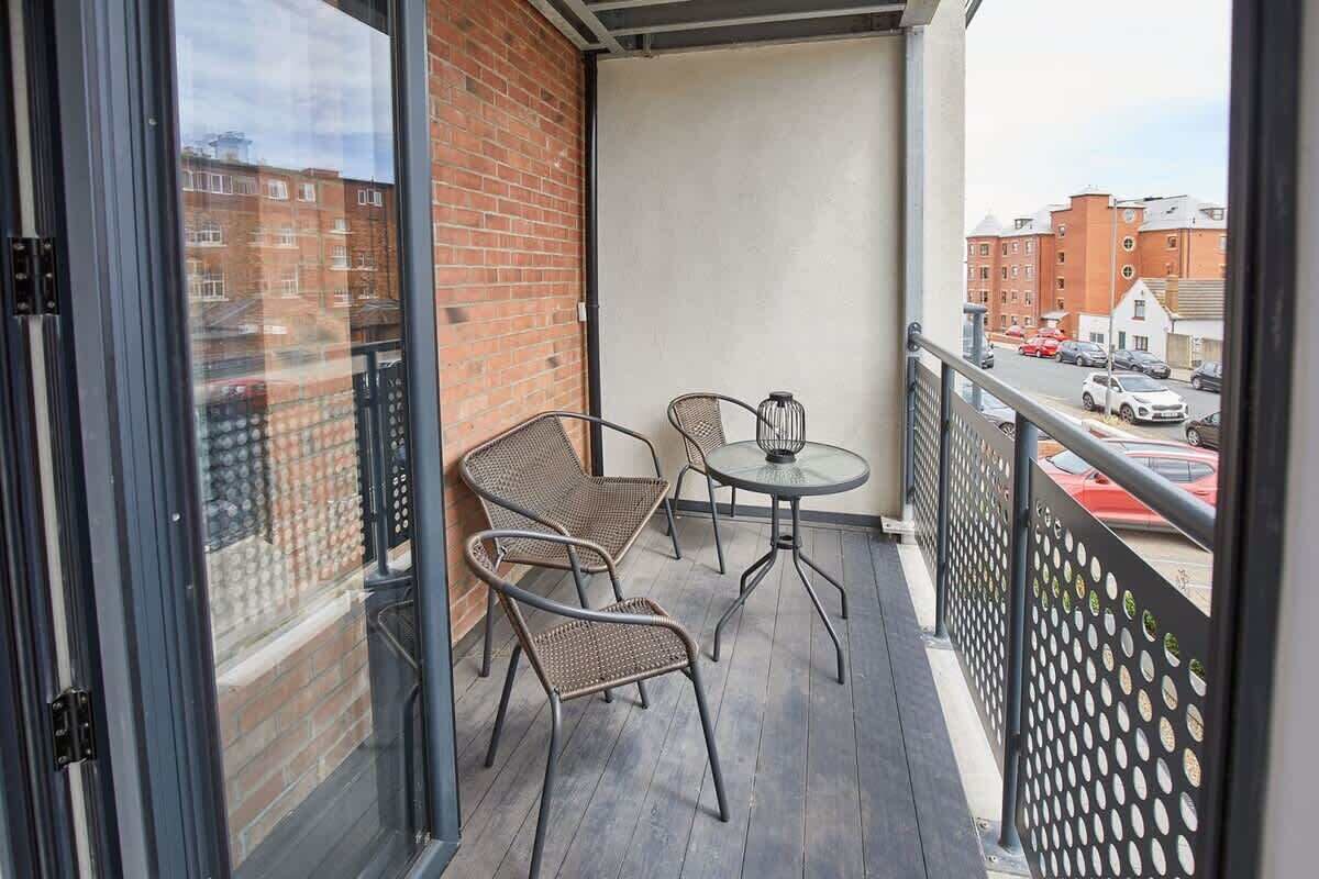 A balcony with two wicker chairs and a small round table, overlooking a street with parked cars and buildings.