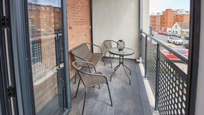 Un balcon avec deux fauteuils en osier et une petite table ronde, donnant sur une rue bordée de voitures garées et d’immeubles.