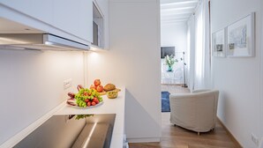 Una cocina moderna con un extractor de acero inoxidable, un tazón de verduras frescas y un comedor con una silla blanca y un jarrón de flores.