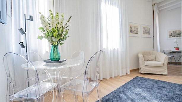 Una sala de estar moderna con una mesa redonda de cristal, sillas de plástico transparente, un jarrón turquesa con flores blancas y una alfombra azul.