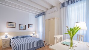 Una habitación de hotel con cama, mesitas de noche, escritorio, silla, lámpara, un jarrón con flores y un armario.