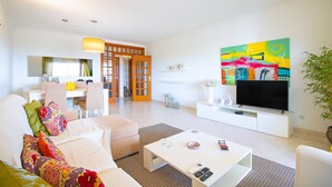 Ein modernes Wohnzimmer mit Sofa, Couchtisch und Fernseher. Im Hintergrund befindet sich ein Essbereich mit Stühlen und einem Tisch.