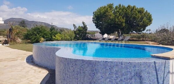 Une piscine ronde, dont le bord est recouvert de carrelage, entourée de chaises longues et d’arbres.