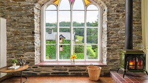 Une pièce aux murs de pierre, dotée d’une grande fenêtre en arc d’où l’on peut admirer un paysage verdoyant, d’une cheminée et d’une petite table sur laquelle est posé un vase de fleurs.