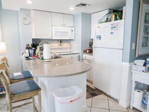 Una cocina con armarios blancos, microondas, estufa y refrigerador. Hay un lavaplatos con grifo, un cubo de basura y una silla.