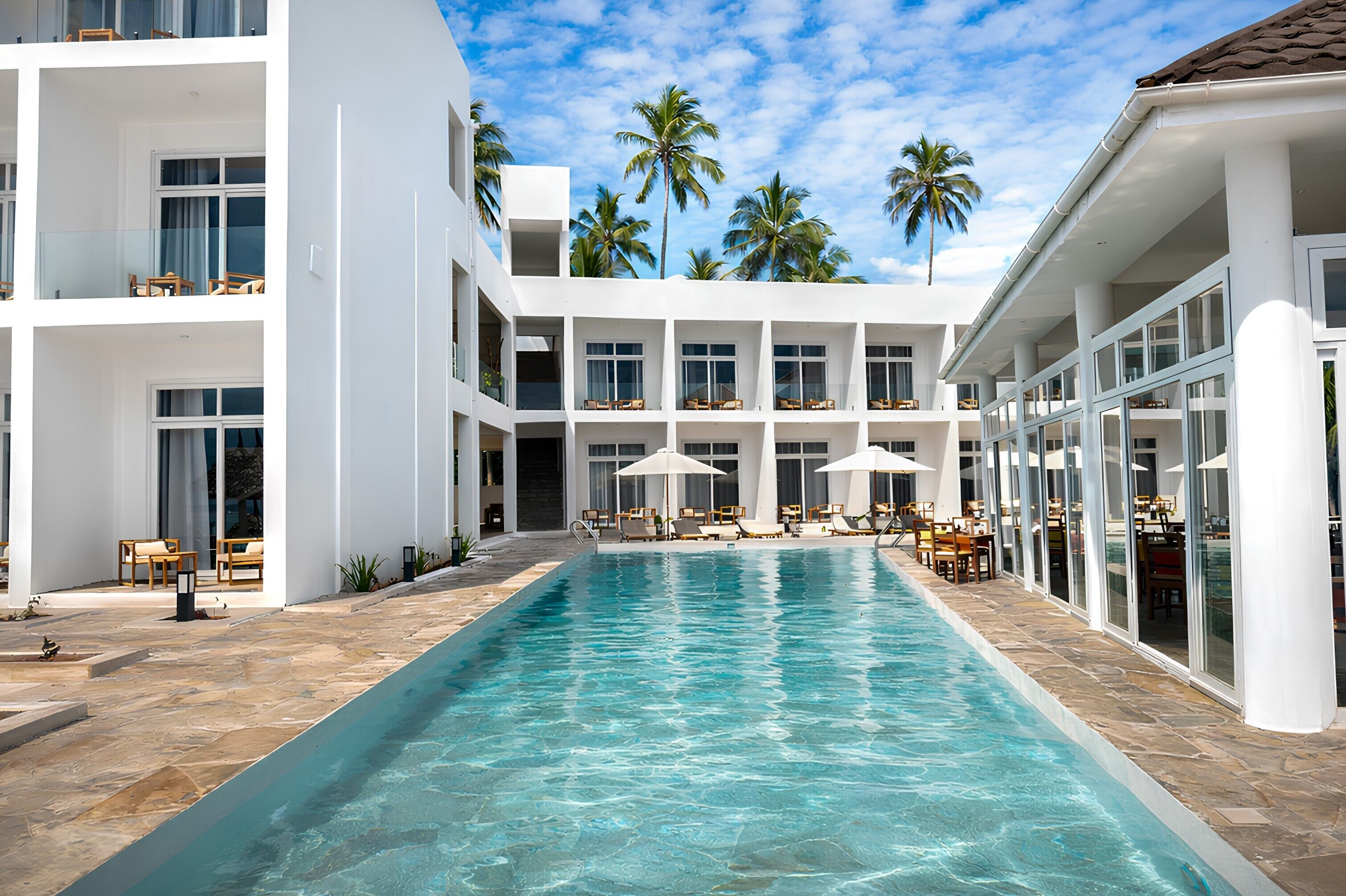 A modern hotel with a central swimming pool, multiple balconies, and palm trees in the background.