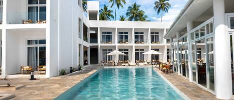 A modern hotel with a central swimming pool, multiple balconies, and palm trees in the background.