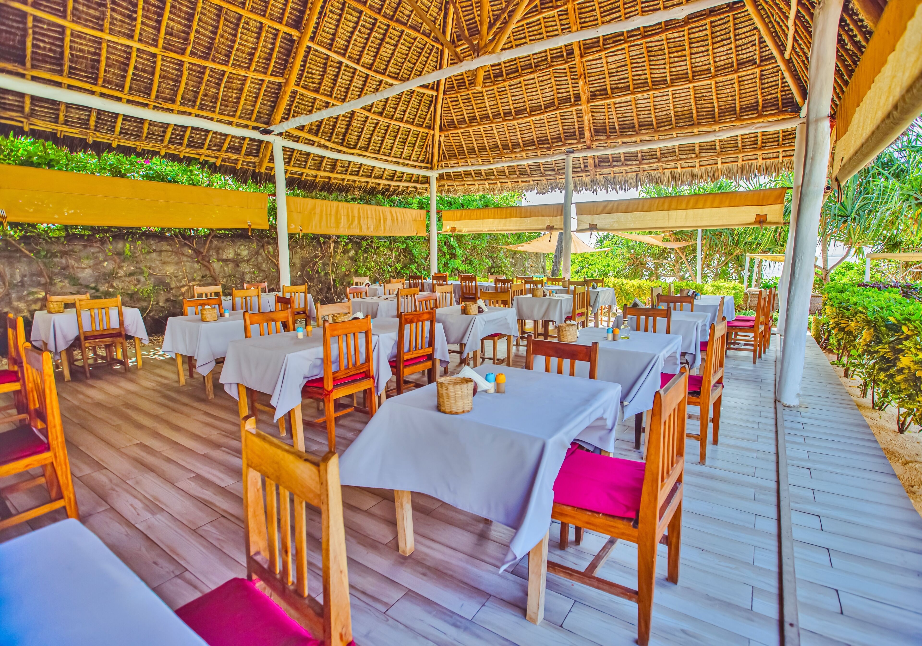 An outdoor dining area with tables and chairs under a thatched roof, surrounded by greenery.