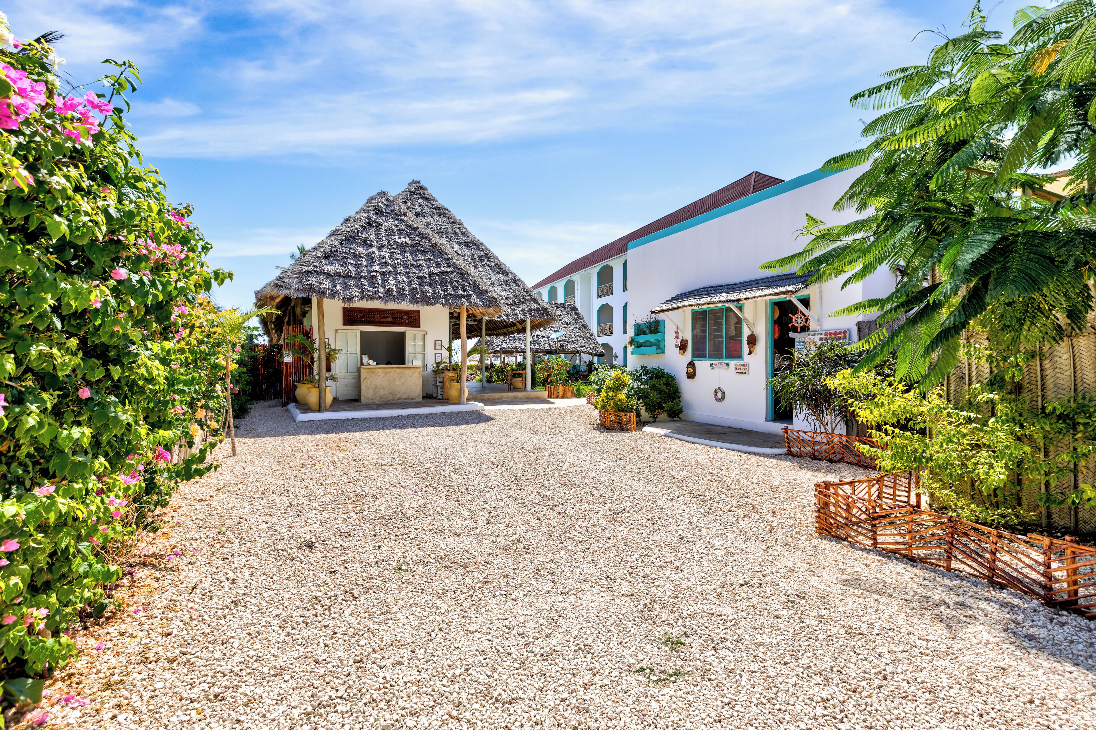 A courtyard with a thatched-roof structure, a white building with a blue roof, and a gravel path.