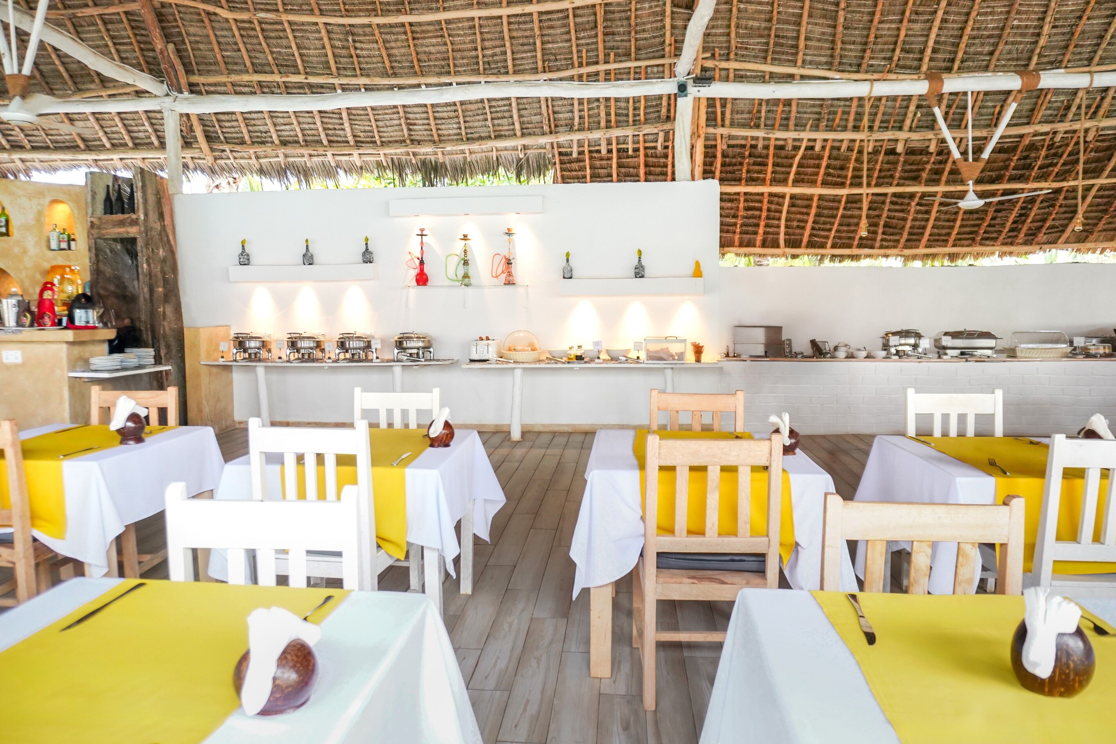 Ein Restaurant mit weißen und gelben Tischtüchern, Holzsesseln und einem Strohdach.