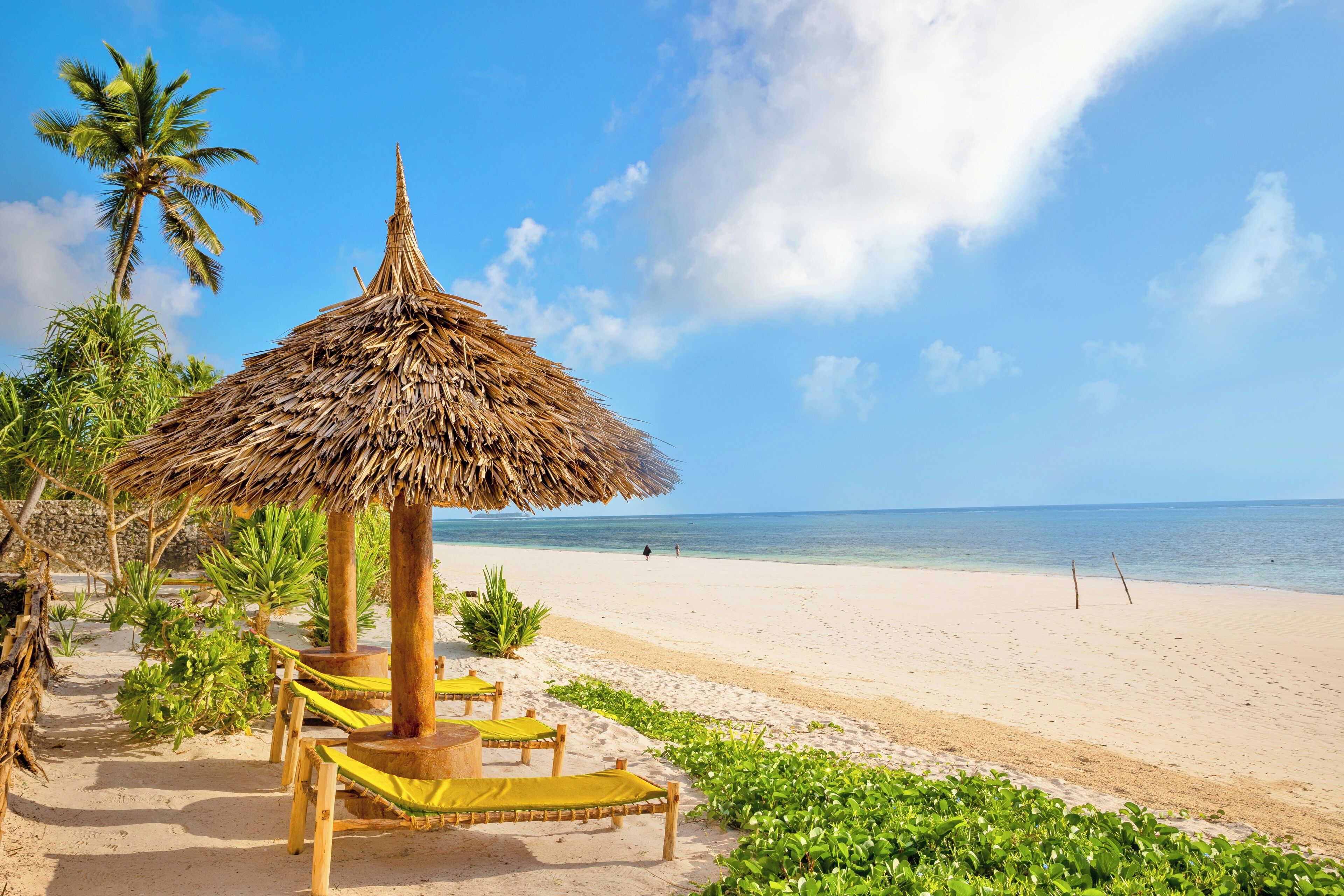 A beach with thatched umbrellas, wooden lounge chairs, and palm trees.