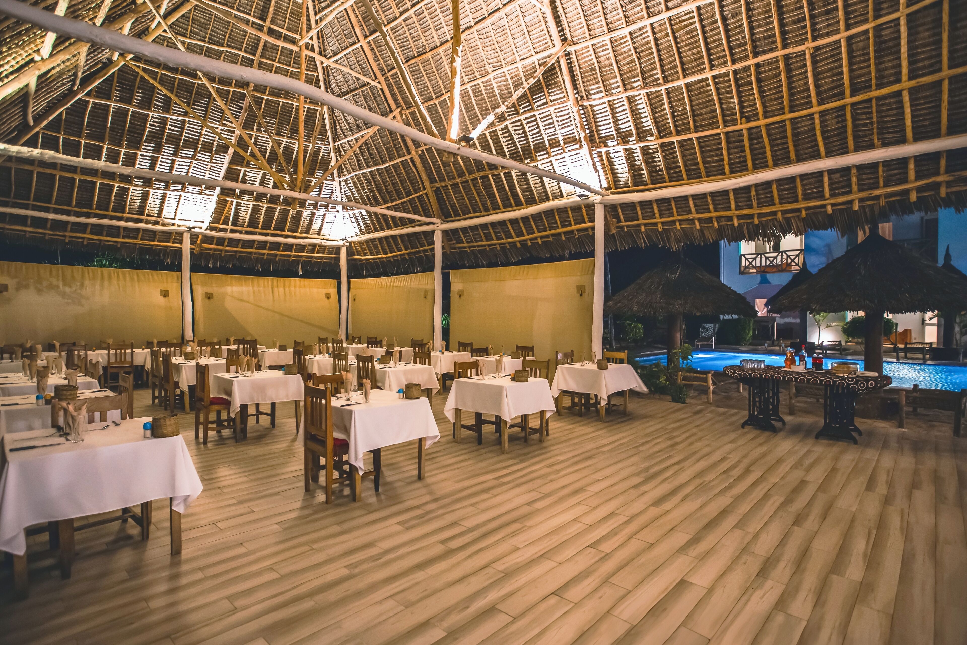 An indoor dining area with tables and chairs, a wooden floor, and a pool in the background.