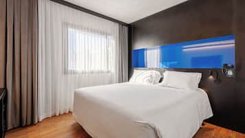 Ein Hotelzimmer mit einem großen Bett, Holzboden, einem Fenster mit durchsichtigen Vorhängen und einem an der Wand befestigten Fernseher.