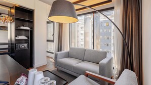 Habitación de hotel moderna con sofá gris, armario negro, ventana grande con cortinas y lámpara colgante.