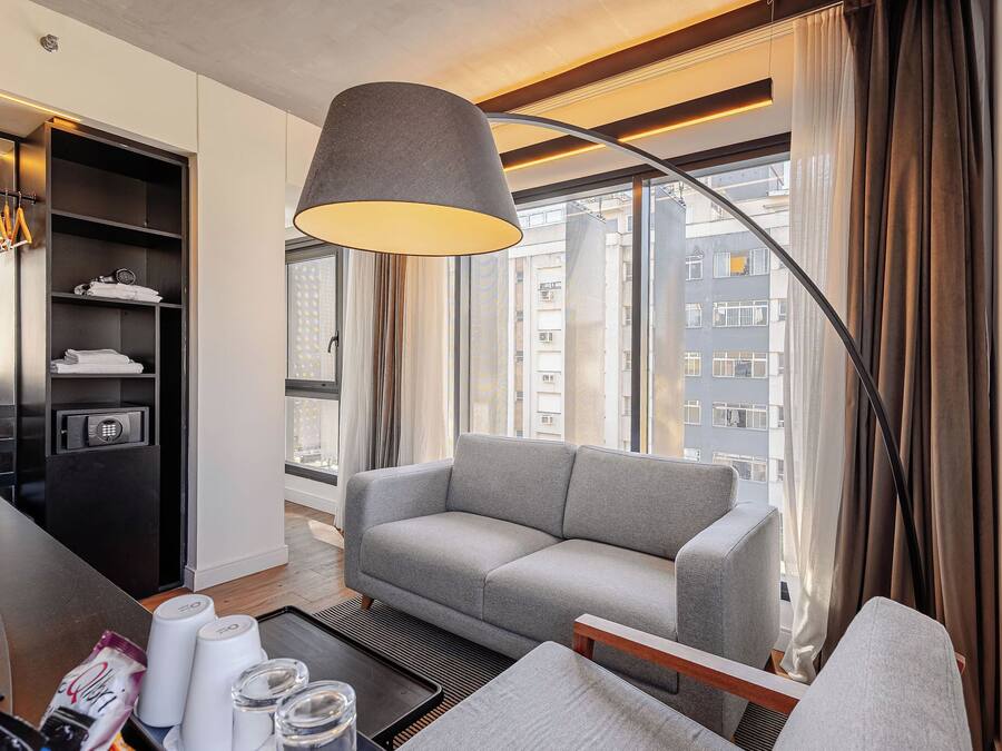 A modern hotel room with a grey sofa, a black wardrobe, a large window with curtains, and a hanging light fixture.