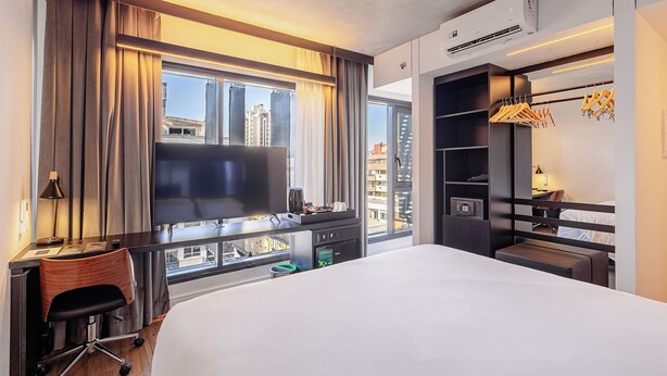 Habitación de hotel con una cama grande, un escritorio con una silla, un televisor, un armario y vistas a la ciudad.