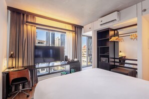Quarto de hotel com uma cama grande, uma mesa com cadeira, televisão, guarda-roupa e vista para a cidade.