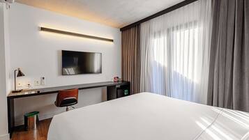 Ein Hotelzimmer mit Bett, Schreibtisch mit Stuhl, Fernseher und einem Fenster mit Vorhängen.