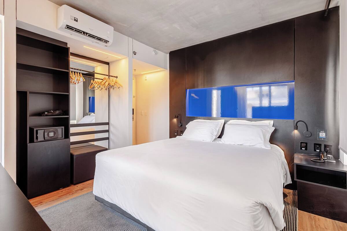A modern hotel room with a large bed, a wardrobe with hanging clothes, a desk with a lamp, and an air conditioning unit.