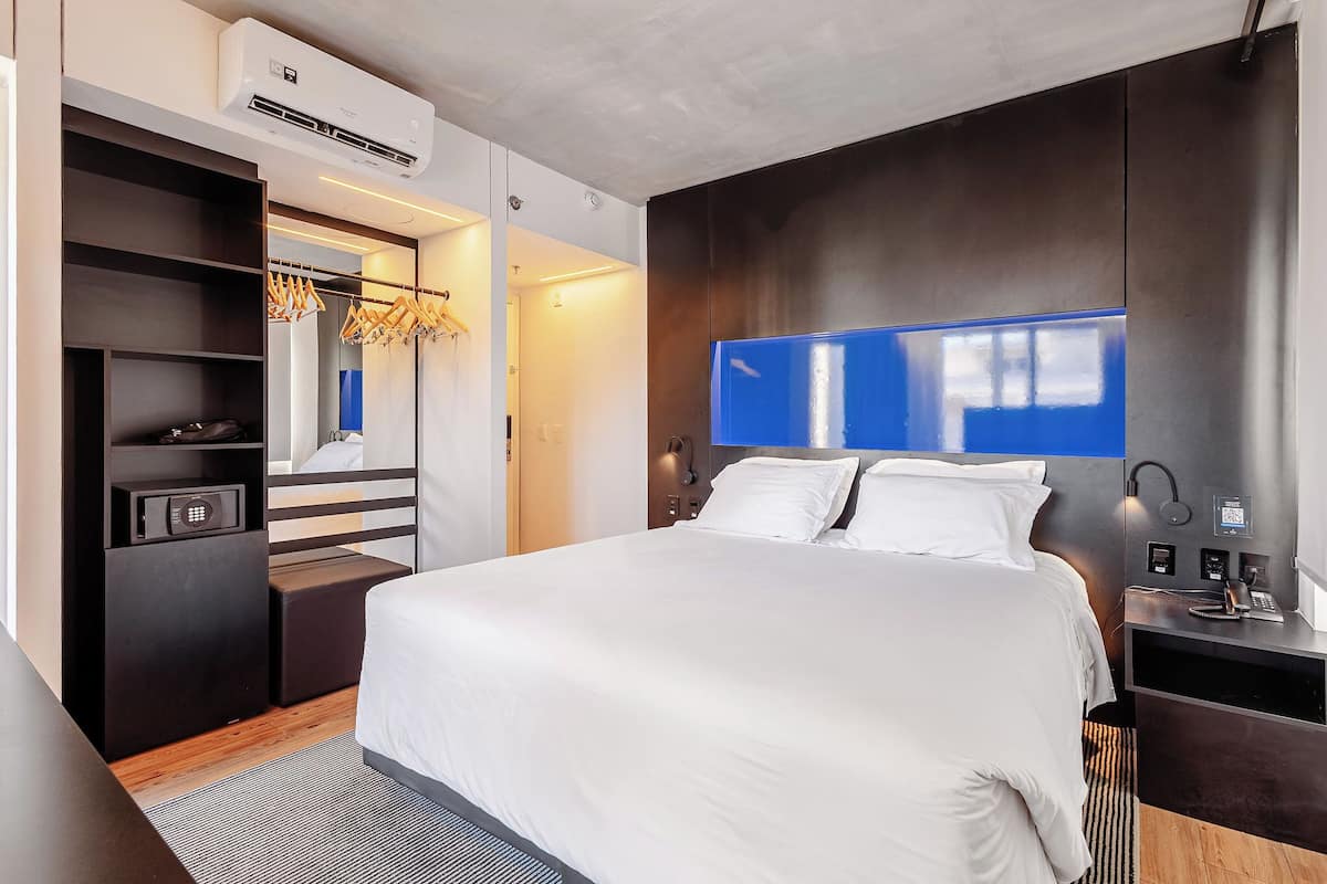 A modern hotel room with a large bed, a wardrobe with hanging clothes, a desk with a lamp, and an air conditioning unit.