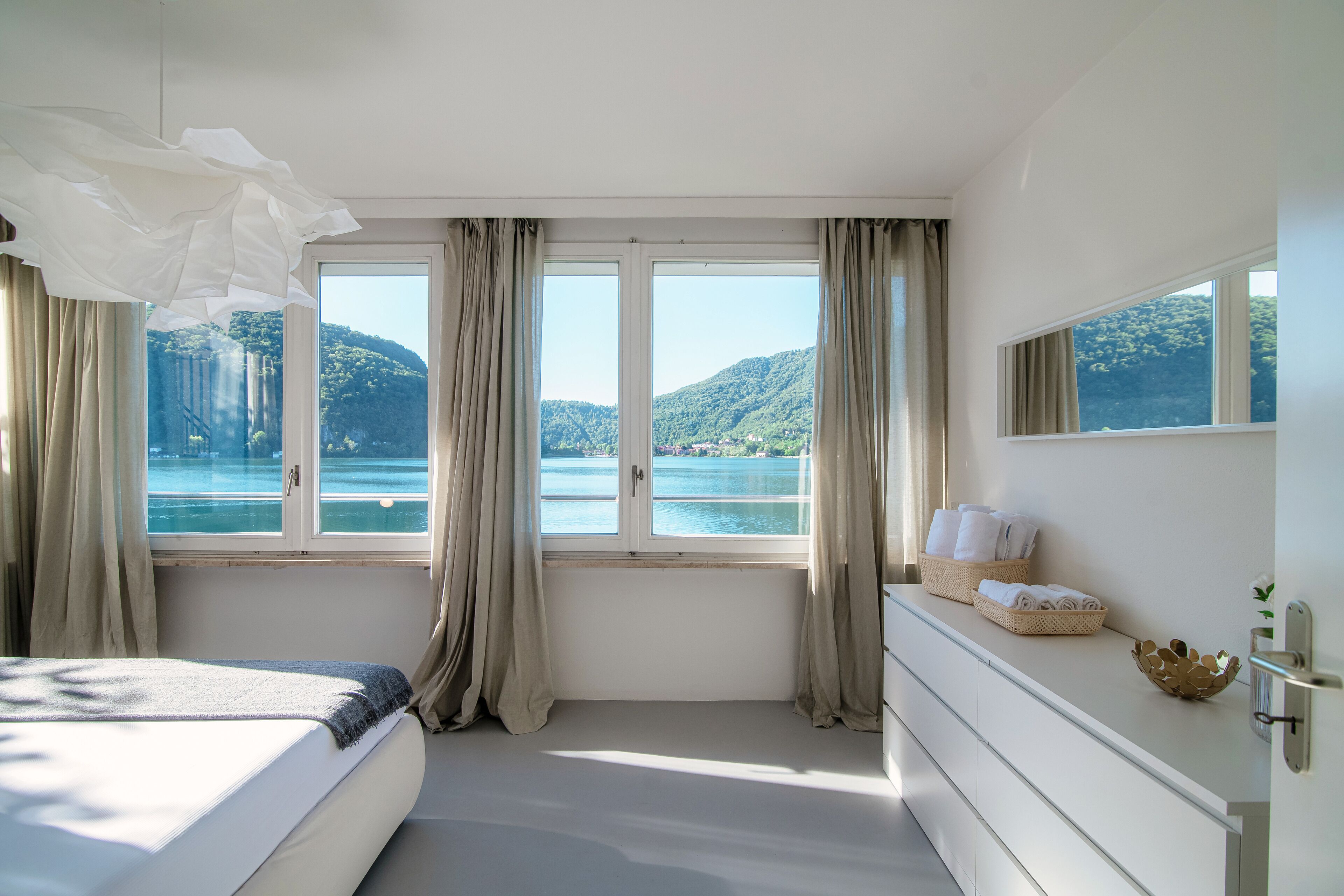 A bedroom with a large window, a bed, a dresser, and a view of a lake and mountains.