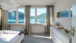 Een slaapkamer met een groot raam, een bed, een kaptafel en uitzicht op een meer en bergen.