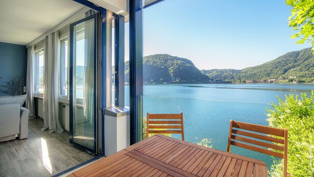 Een balkon met een houten tafel en stoelen, met uitzicht op een meer met bergen op de achtergrond.