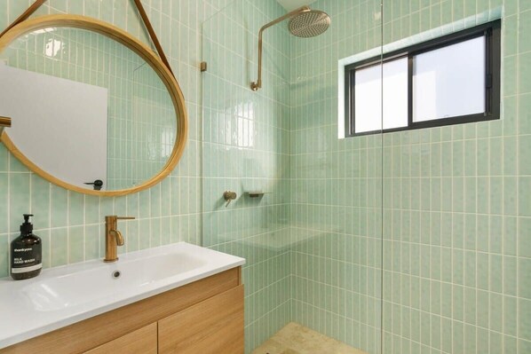 A bathroom with a sink, mirror, shower, and a window.