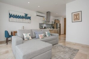 A modern living room with a grey sectional sofa, a small table, and a blue chair. A kitchen with a tiled backsplash and stainless steel appliances is visible in the background.
