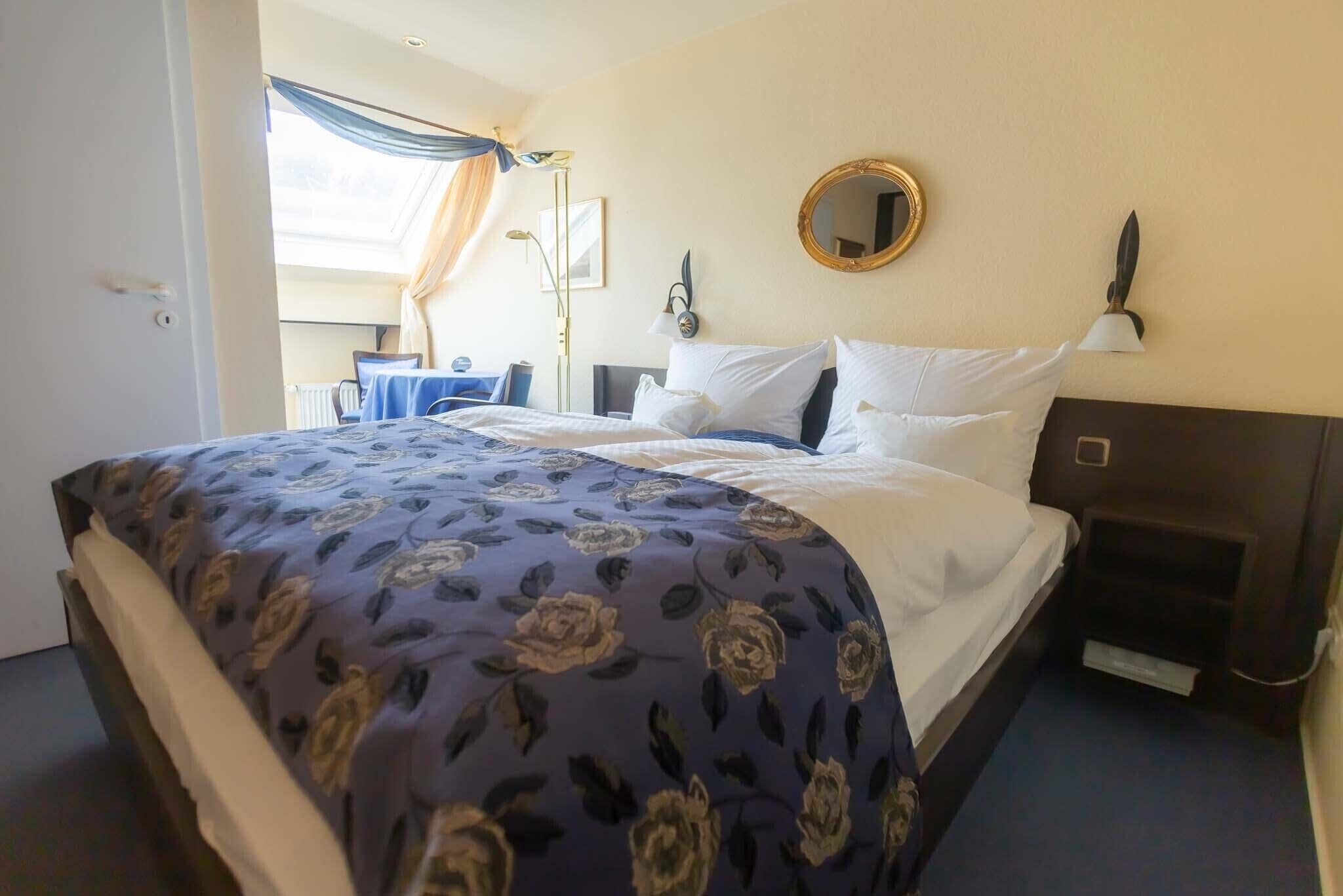 Une chambre d’hôtel avec un lit, une table de chevet, une lampe, un miroir et une fenêtre avec des rideaux.