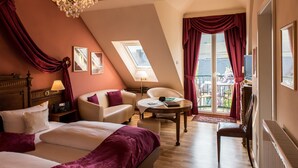 Quarto de hotel com uma cama grande, um sofá, uma cadeira, uma mesa pequena, um candelabro e uma varanda com cortinas.