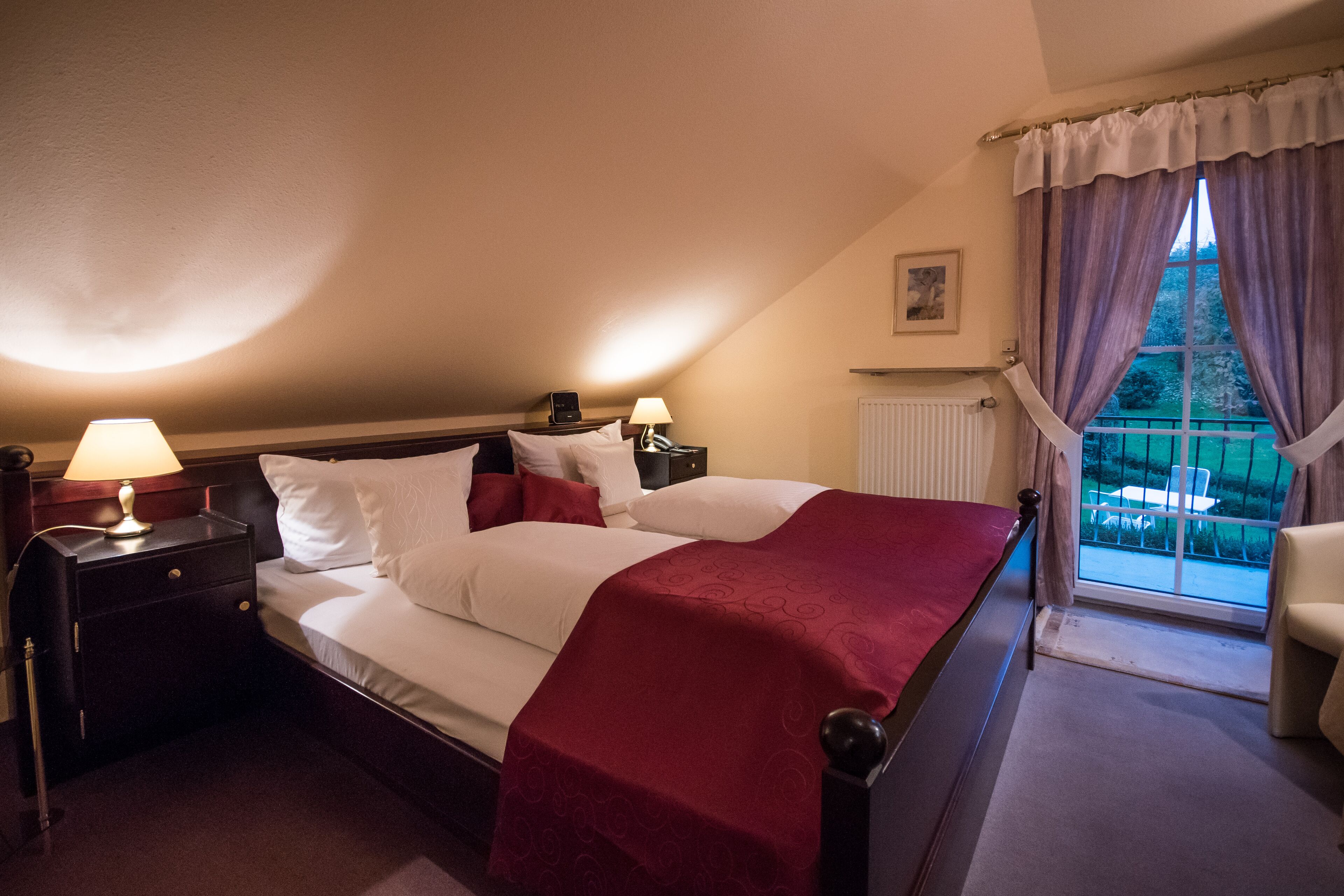 A bedroom with a large bed, two bedside tables with lamps, a window with curtains, and a view of the outdoors.